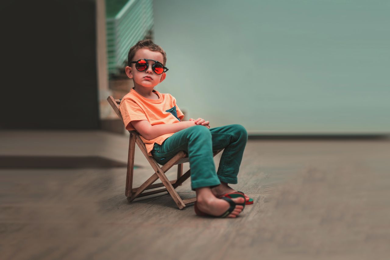 Young boy in sunglasses and vibrant outfit sitting confidently on a wooden chair.